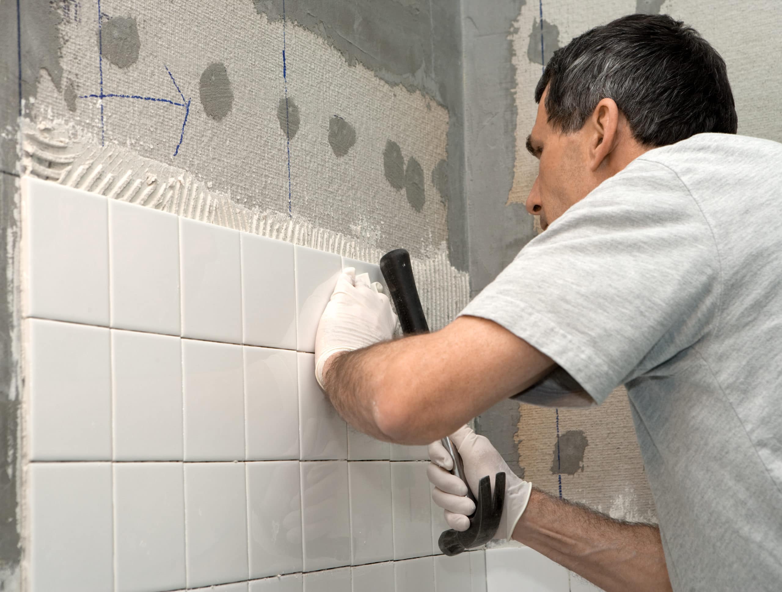 Man setting tile on cement board. He is tapping the tile in place with the handle of a hammer.
